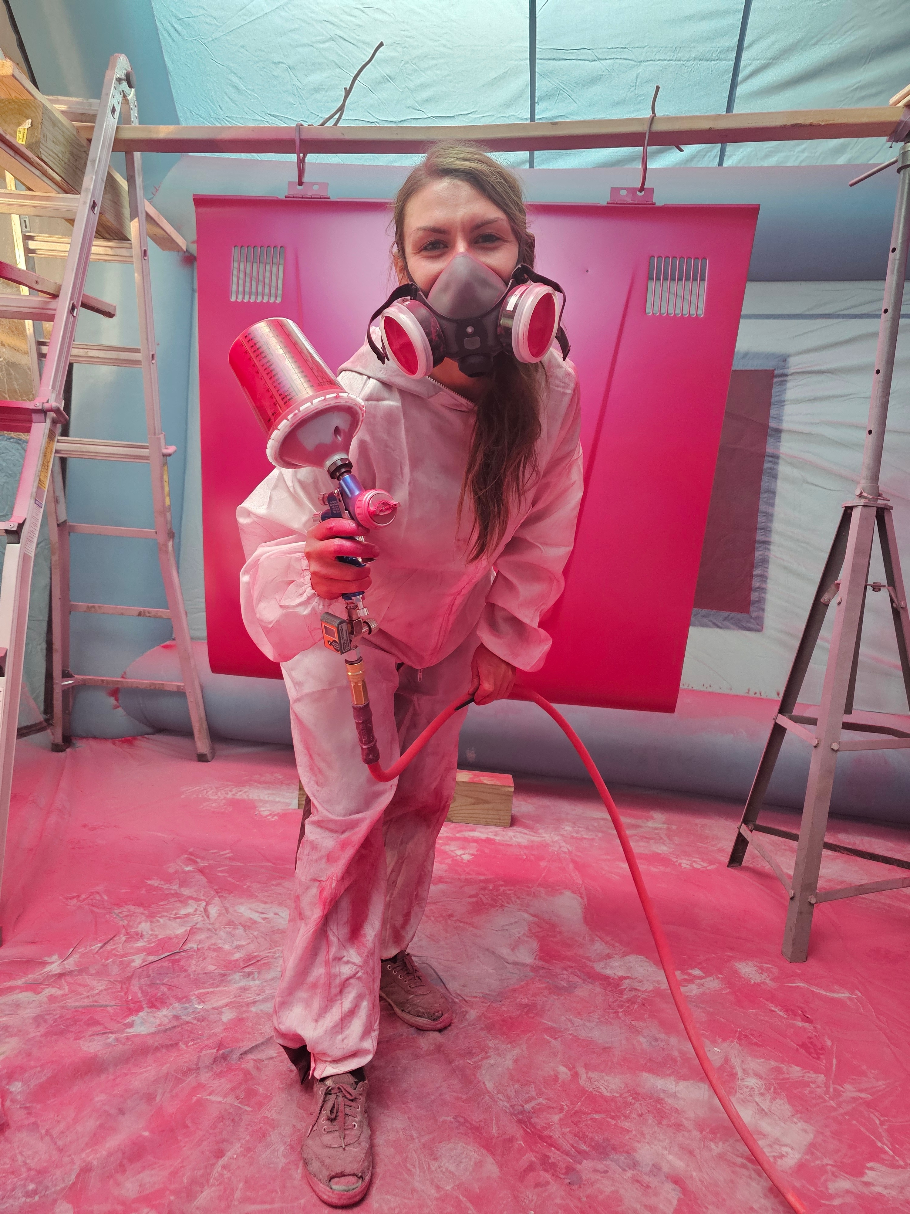 A woman wearing a protective suit holds a paint sprayer and is covered in hot pink paint.